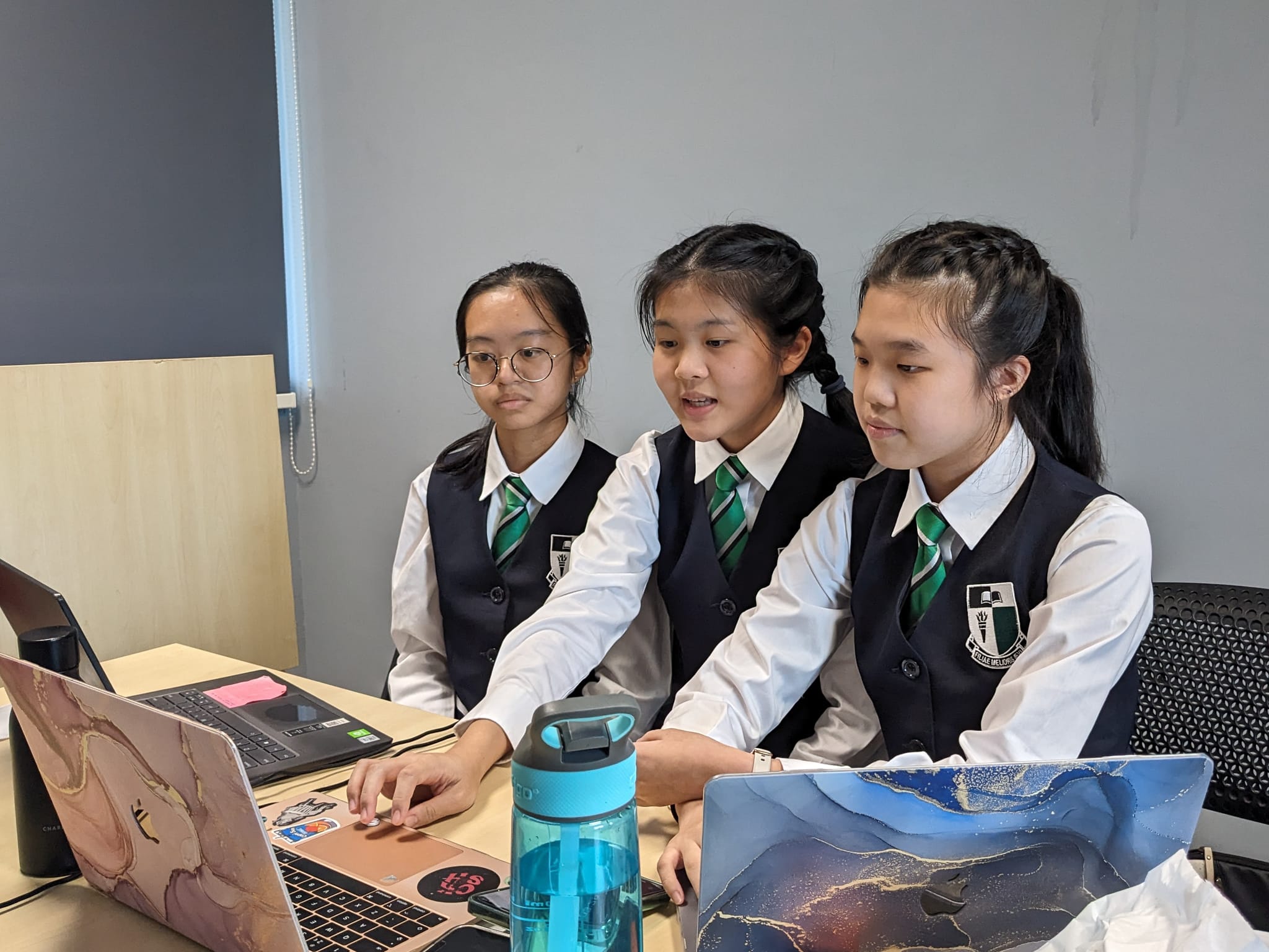 Three uniformed schoolgirls in dark vests and green ties sit together at a desk looking at a laptop.